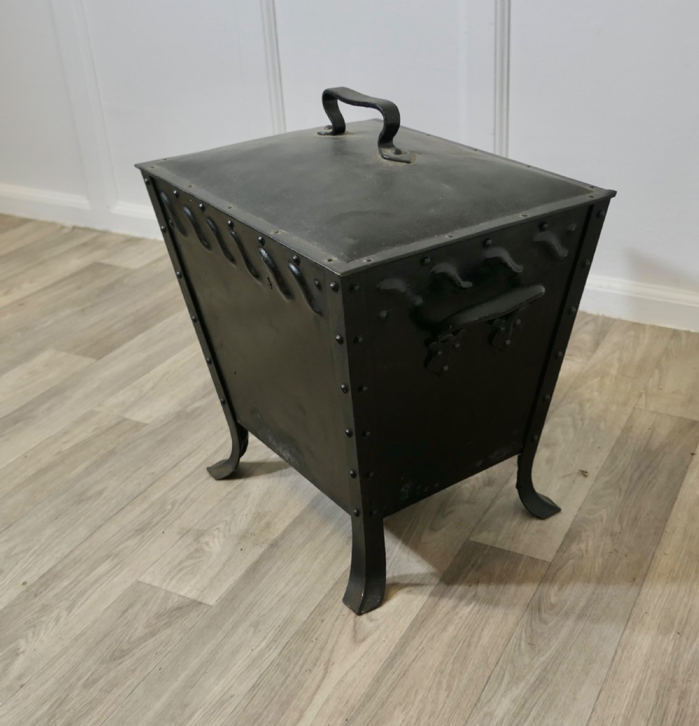 A black riveted metal Arts and Crafts coal box featuring a domed lid with a central handle, decorative wave detailing, and splayed metal legs. View V