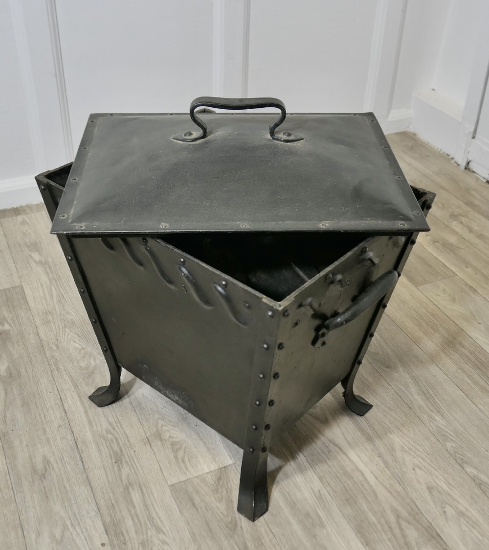A black riveted metal Arts and Crafts coal box featuring a domed lid with a central handle, decorative wave detailing, and splayed metal legs. View VI