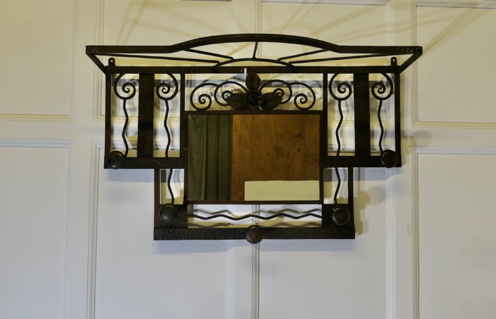 A French Art Deco dark wrought iron wall-mounted rack featuring a central rectangular mirror, top hat shelf, decorative scrollwork, and five coat pegs.