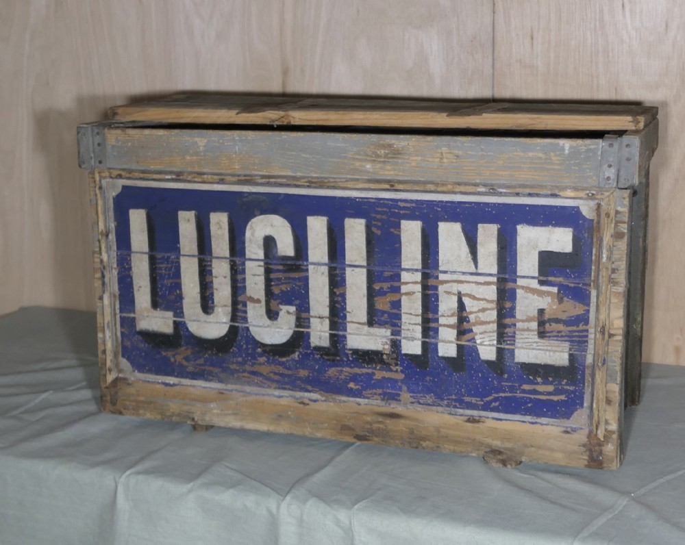A rectangular weathered wooden shipping crate featuring painted blue typography spelling Luciline across the front panel with metal edge reinforcements.