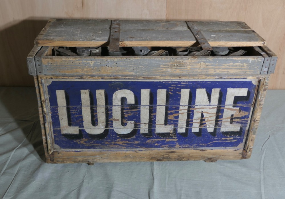 A rectangular weathered wooden shipping crate featuring painted blue typography spelling Luciline across the front panel with metal edge reinforcements. View II
