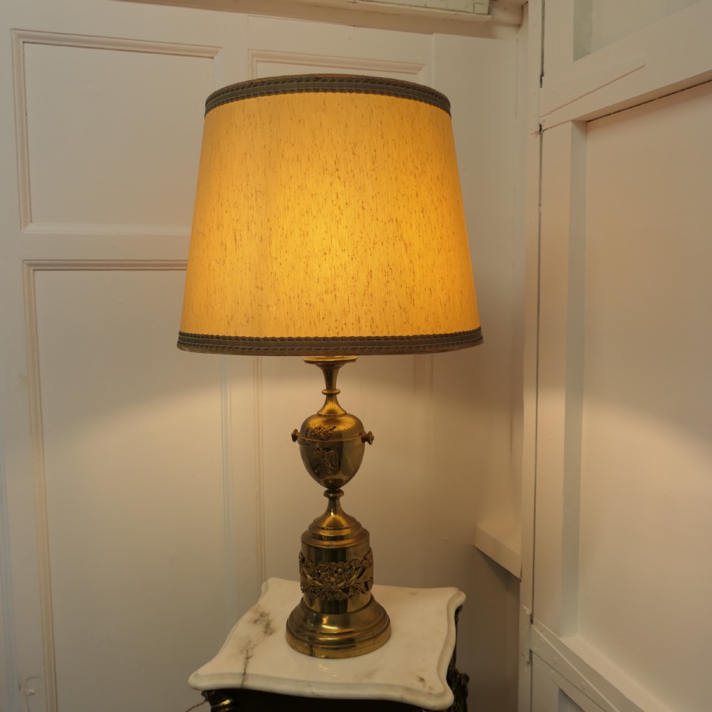 An antique Neoclassical brass table lamp featuring an urn-shaped stem, classical mask reliefs around the base, and a gold fabric shade.