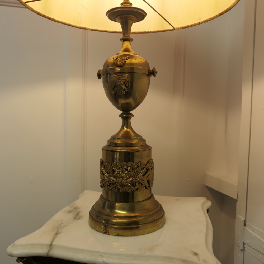 An antique Neoclassical brass table lamp featuring an urn-shaped stem, classical mask reliefs around the base, and a gold fabric shade. View V