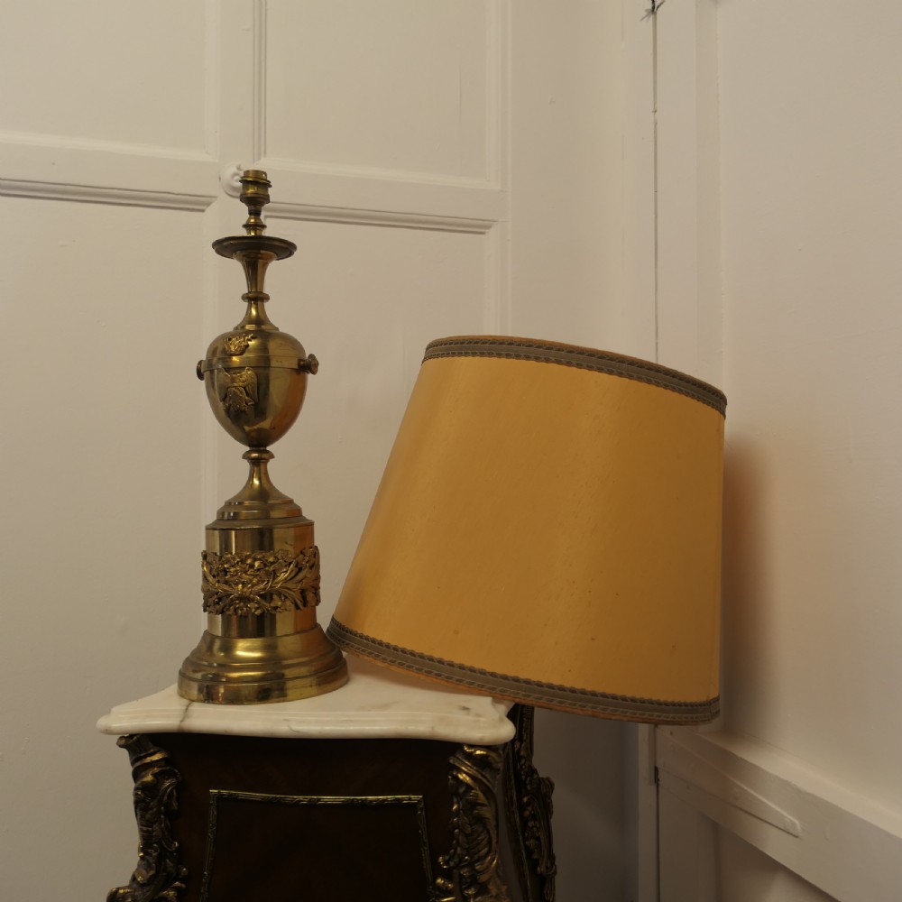 An antique Neoclassical brass table lamp featuring an urn-shaped stem, classical mask reliefs around the base, and a gold fabric shade. View VII