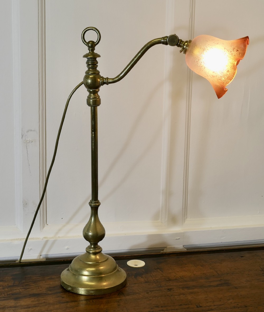 An antique brass desk lamp featuring a weighted circular base, an adjustable curved arm, and an etched pinkish-amber glass shade.
