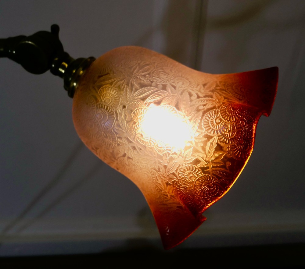 An antique brass desk lamp featuring a weighted circular base, an adjustable curved arm, and an etched pinkish-amber glass shade. View III