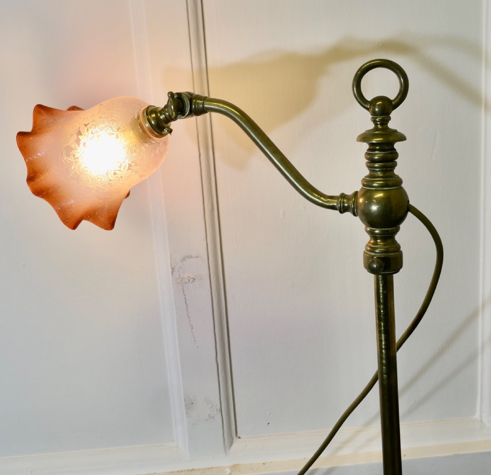 An antique brass desk lamp featuring a weighted circular base, an adjustable curved arm, and an etched pinkish-amber glass shade. View VII