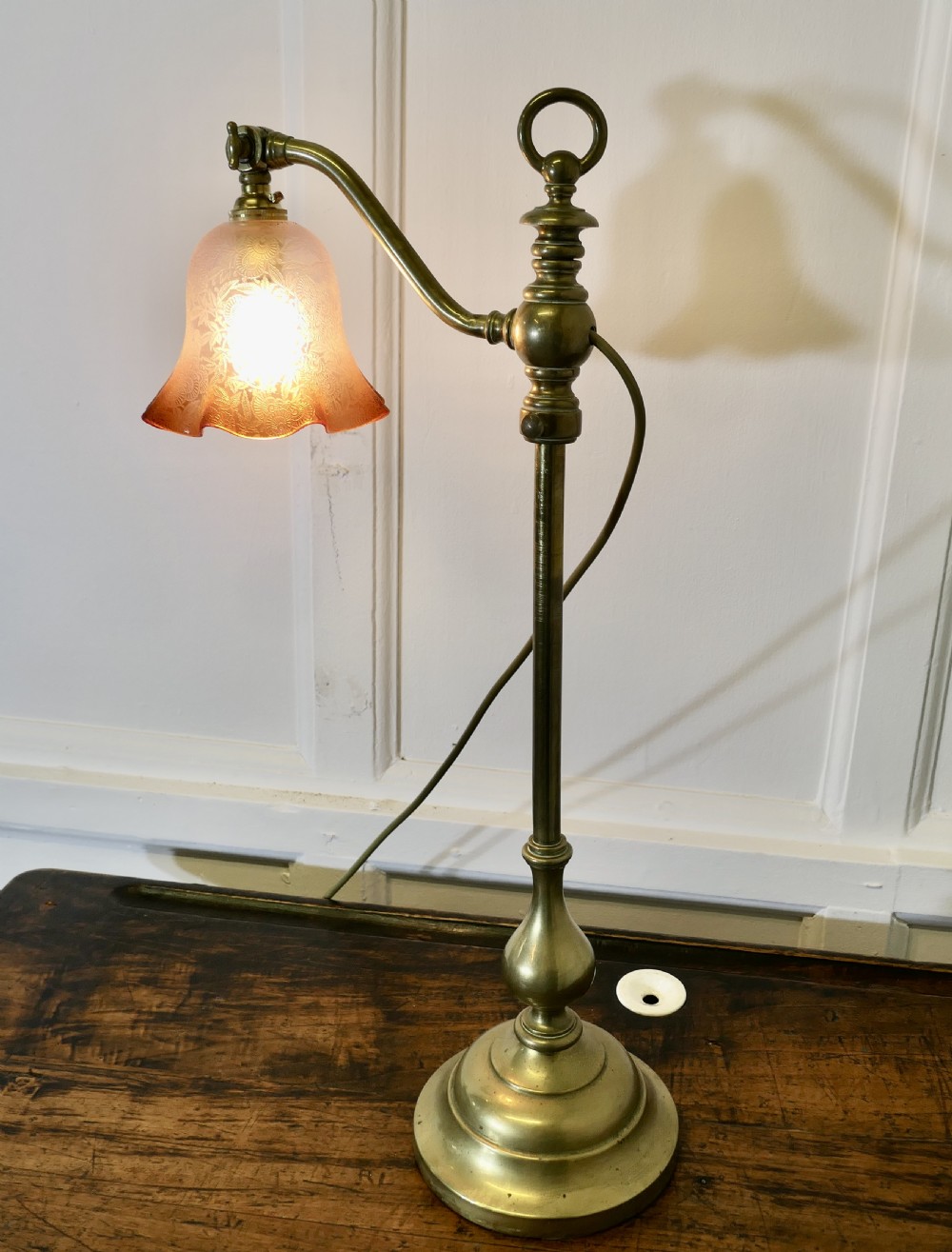 An antique brass desk lamp featuring a weighted circular base, an adjustable curved arm, and an etched pinkish-amber glass shade. View IX