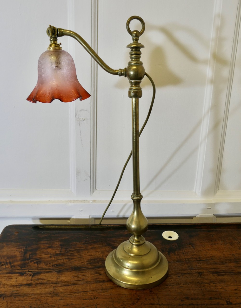 An antique brass desk lamp featuring a weighted circular base, an adjustable curved arm, and an etched pinkish-amber glass shade. View X