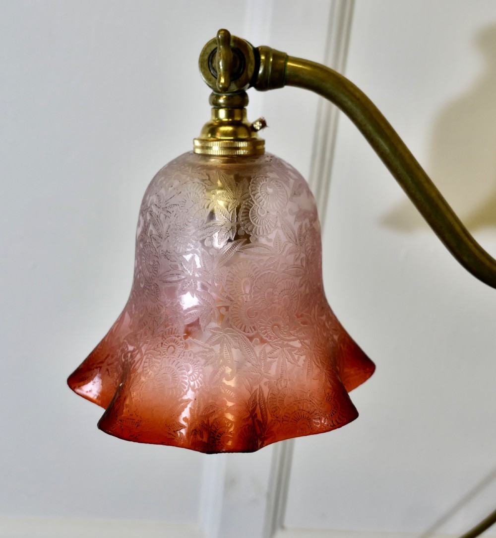 An antique brass desk lamp featuring a weighted circular base, an adjustable curved arm, and an etched pinkish-amber glass shade. View XI