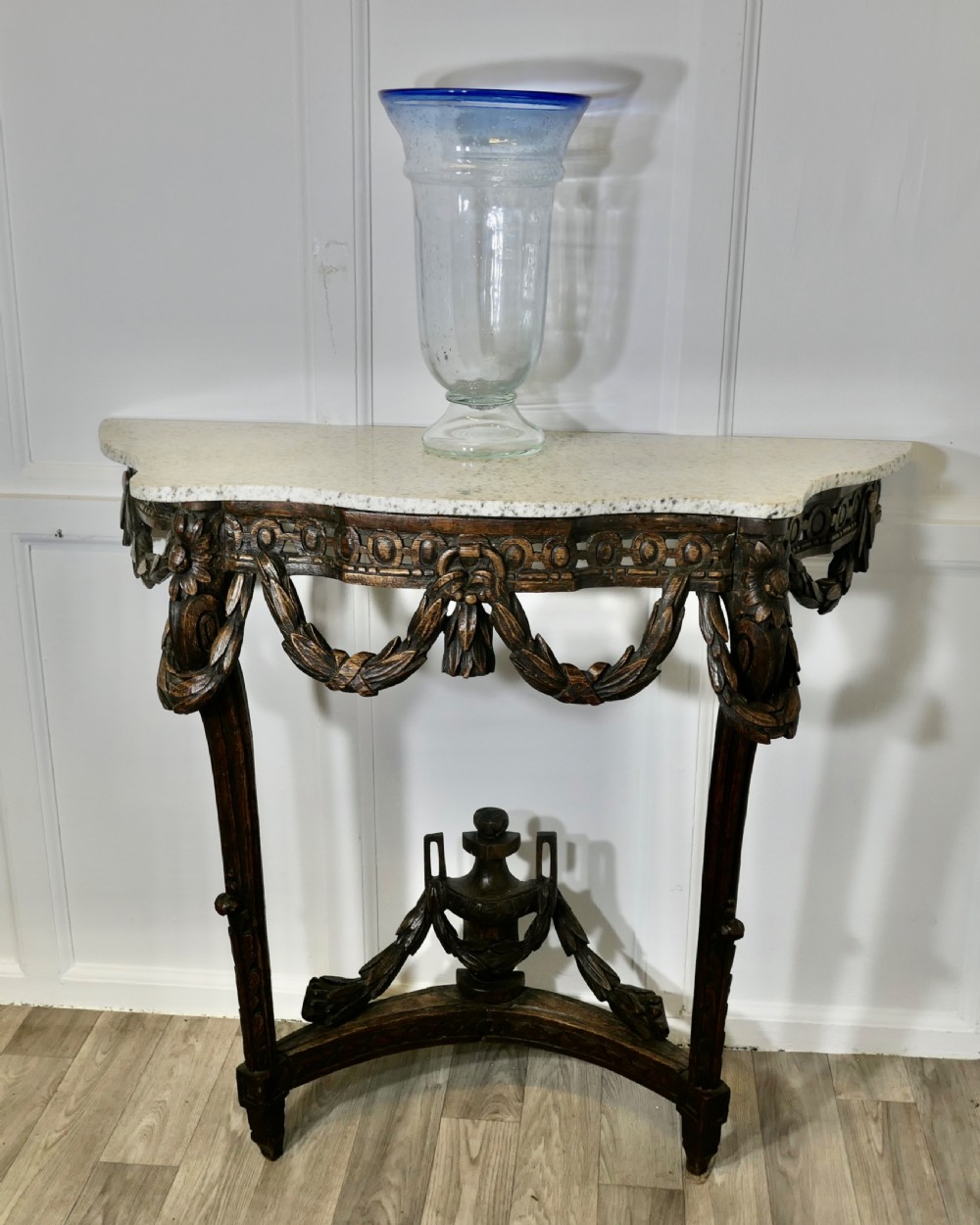 A dark hand-carved wooden French Neoclassical console table featuring drape festoons, a guilloche apron, a curved stretcher with an urn finial, and a light granite top. View IV