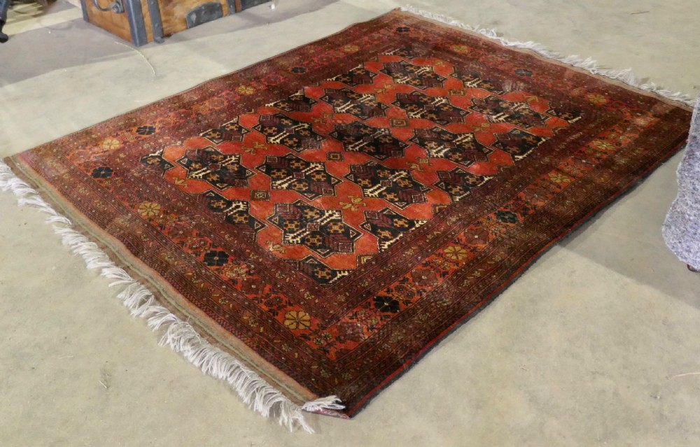 A rectangular vintage wool area rug featuring dark brown and navy geometric motifs on a terracotta red field with patterned borders and fringe.