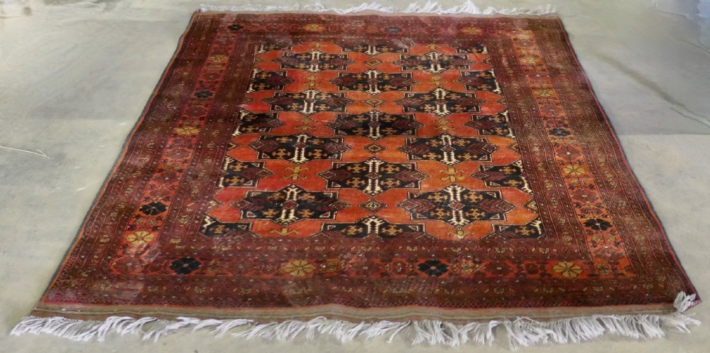 A rectangular vintage wool area rug featuring dark brown and navy geometric motifs on a terracotta red field with patterned borders and fringe. View VI