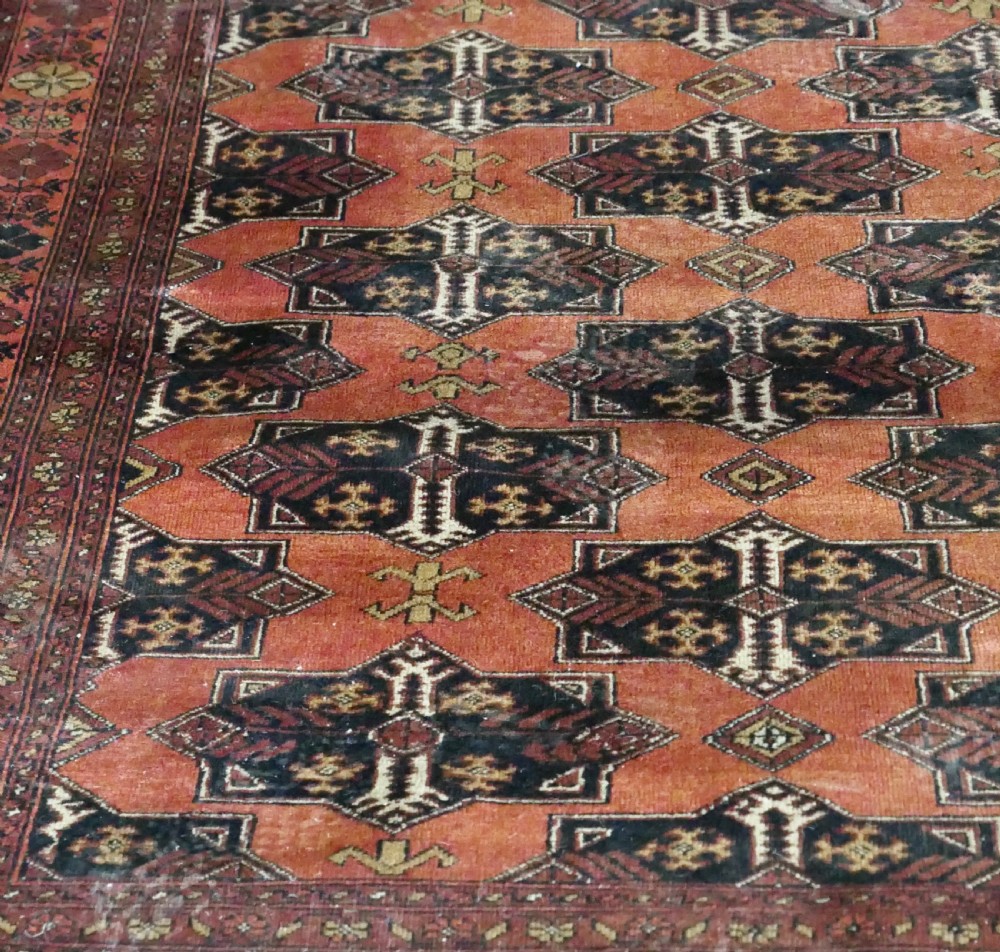 A rectangular vintage wool area rug featuring dark brown and navy geometric motifs on a terracotta red field with patterned borders and fringe. View X