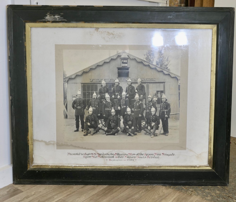 A large Edwardian framed photograph showing a posed group of firefighters in uniform in front of a fire station with a calligraphic inscription along the bottom.