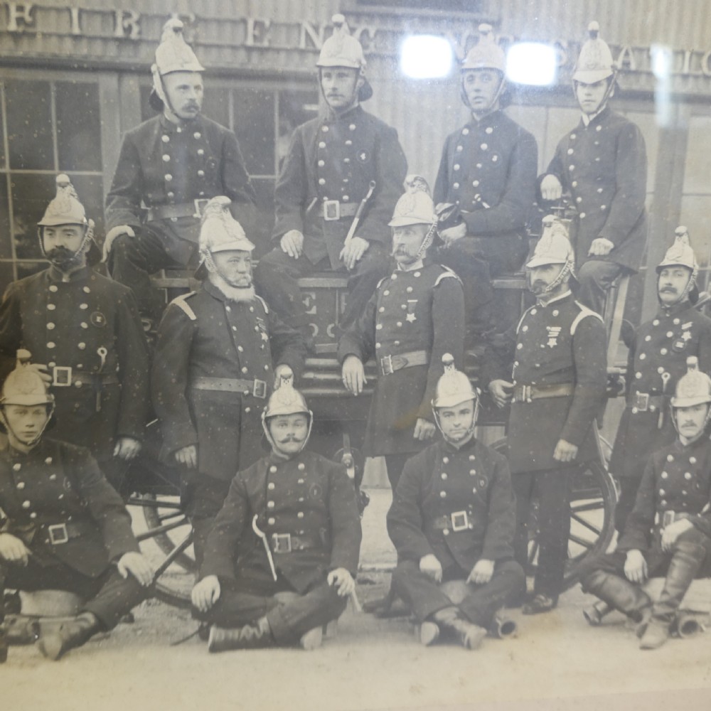 A large Edwardian framed photograph showing a posed group of firefighters in uniform in front of a fire station with a calligraphic inscription along the bottom. View III