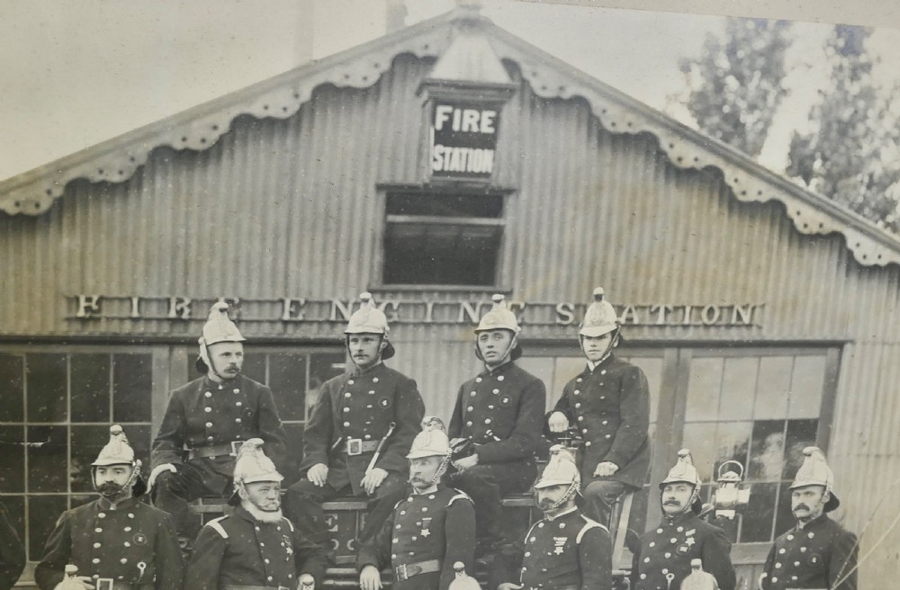 A large Edwardian framed photograph showing a posed group of firefighters in uniform in front of a fire station with a calligraphic inscription along the bottom. View IV