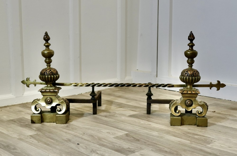 A pair of ornate French Baroque style brass andirons featuring flame finials, pierced spherical sections, scroll bases, and an adjustable twisted brass bar with fleur-de-lis ends.