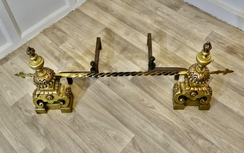 A pair of ornate French Baroque style brass andirons featuring flame finials, pierced spherical sections, scroll bases, and an adjustable twisted brass bar with fleur-de-lis ends. View III