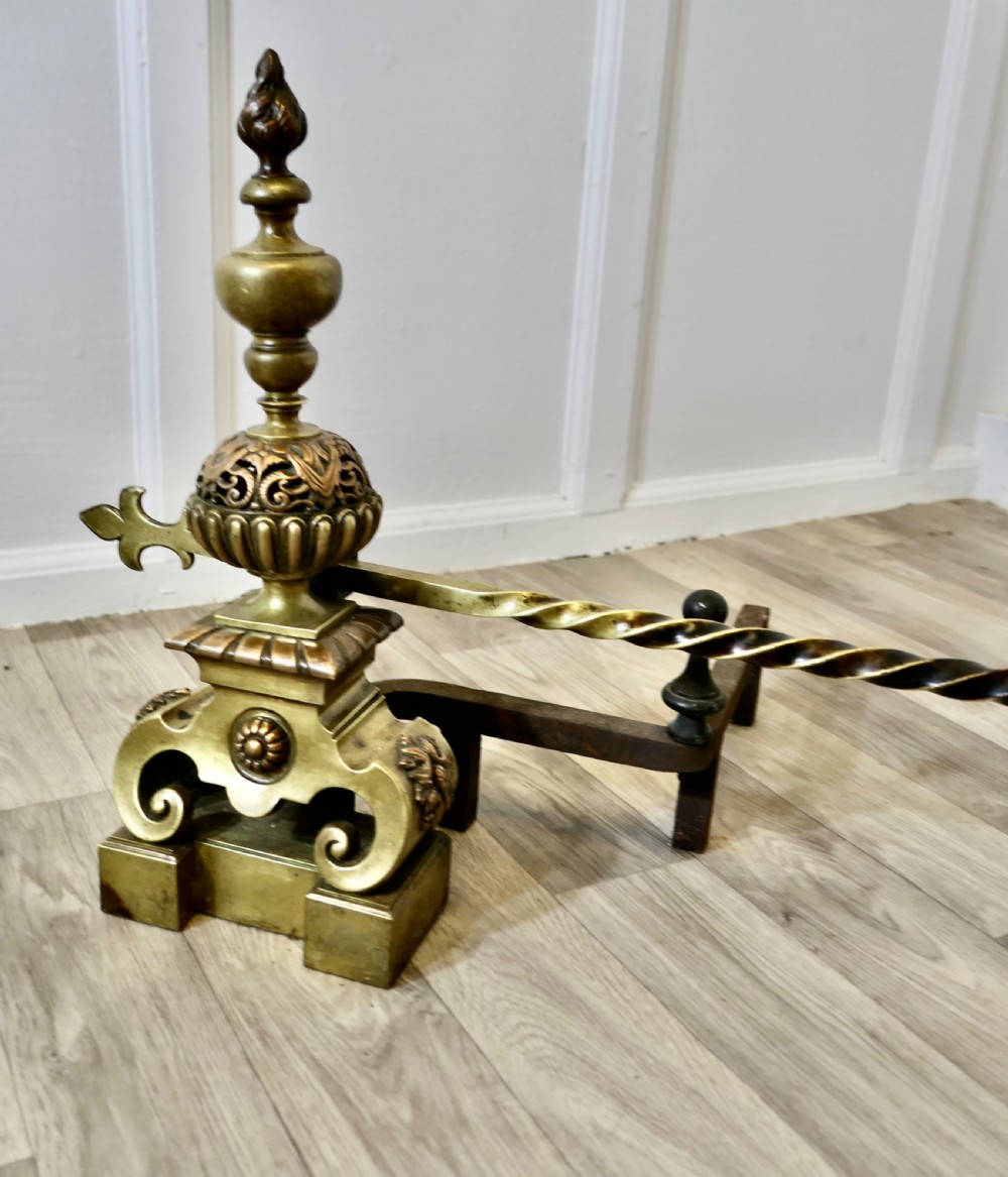 A pair of ornate French Baroque style brass andirons featuring flame finials, pierced spherical sections, scroll bases, and an adjustable twisted brass bar with fleur-de-lis ends. View IV