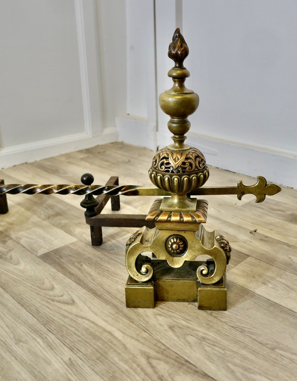 A pair of ornate French Baroque style brass andirons featuring flame finials, pierced spherical sections, scroll bases, and an adjustable twisted brass bar with fleur-de-lis ends. View V