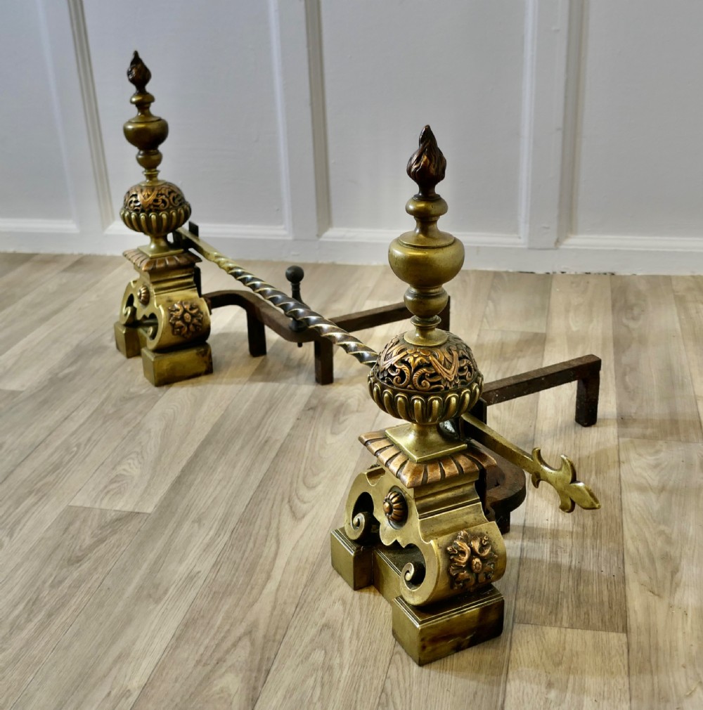 A pair of ornate French Baroque style brass andirons featuring flame finials, pierced spherical sections, scroll bases, and an adjustable twisted brass bar with fleur-de-lis ends. View VI