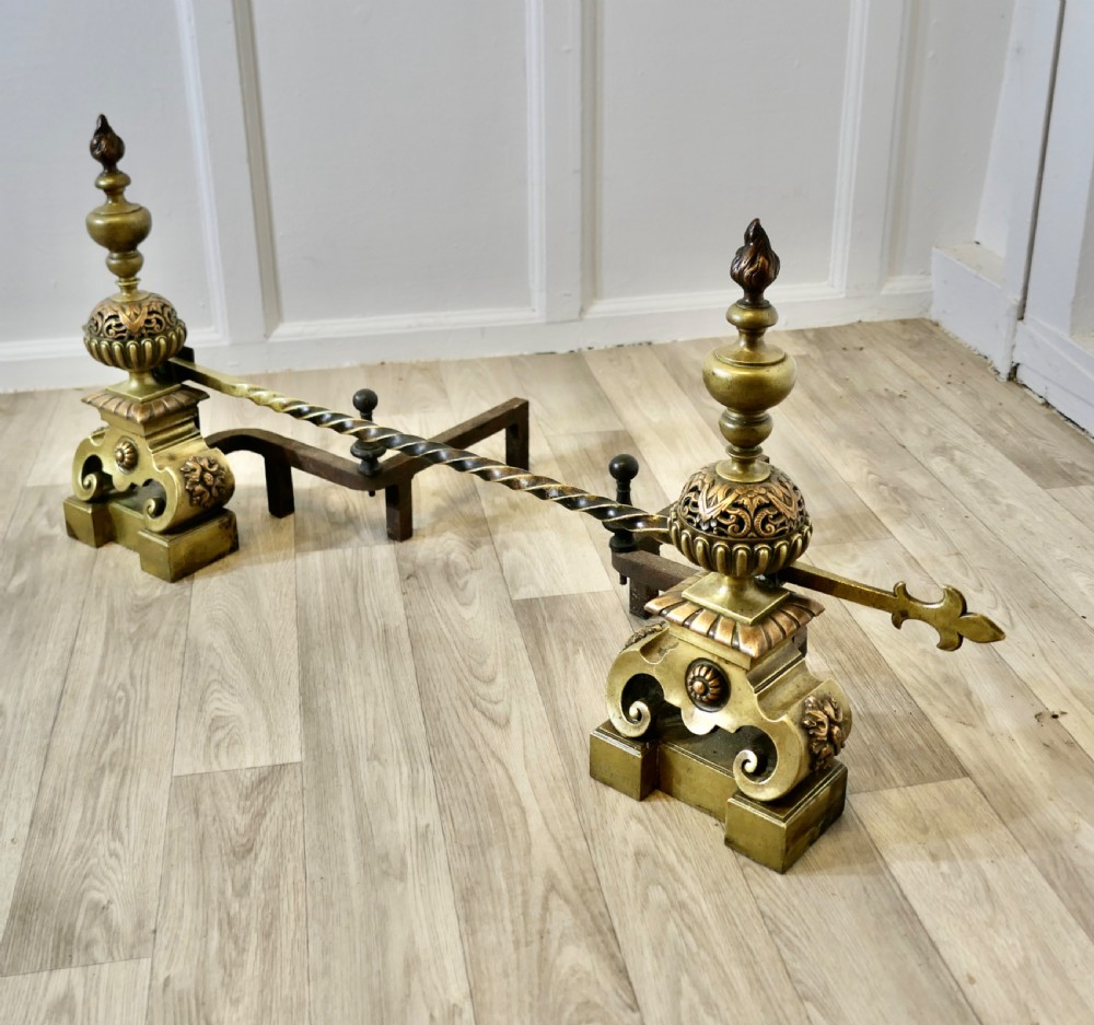 A pair of ornate French Baroque style brass andirons featuring flame finials, pierced spherical sections, scroll bases, and an adjustable twisted brass bar with fleur-de-lis ends. View VII