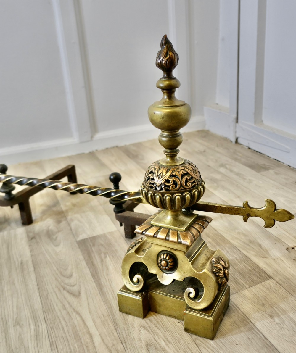 A pair of ornate French Baroque style brass andirons featuring flame finials, pierced spherical sections, scroll bases, and an adjustable twisted brass bar with fleur-de-lis ends. View VIII