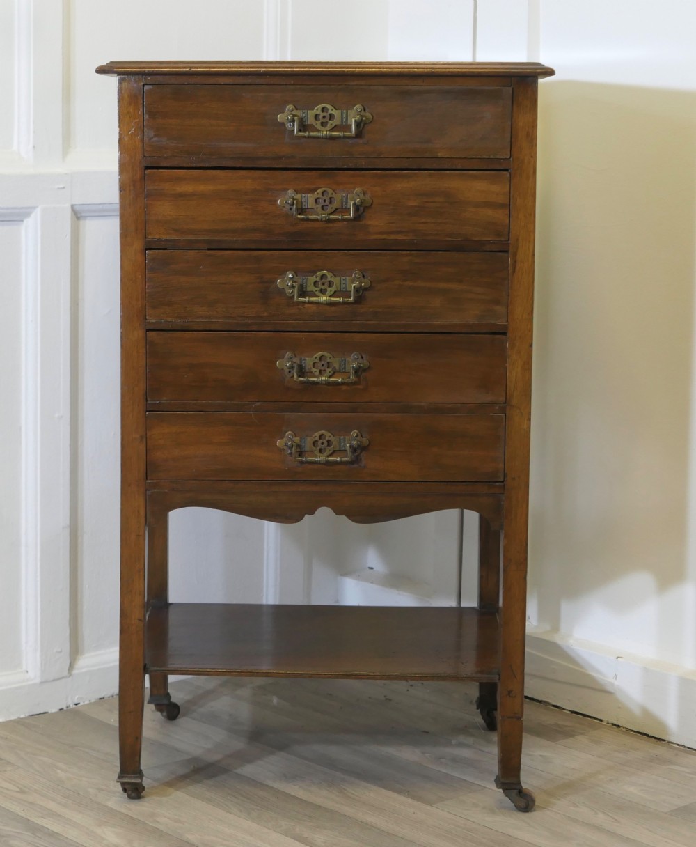 An Arts and Crafts warm brown wood music cabinet with five drawers featuring ornamental brass drop handles, raised on tapered legs with castors and a lower shelf.