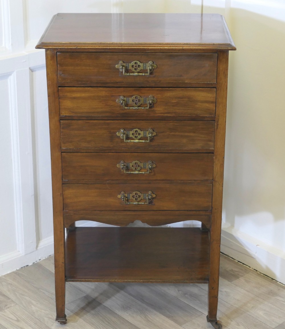 An Arts and Crafts warm brown wood music cabinet with five drawers featuring ornamental brass drop handles, raised on tapered legs with castors and a lower shelf. View II