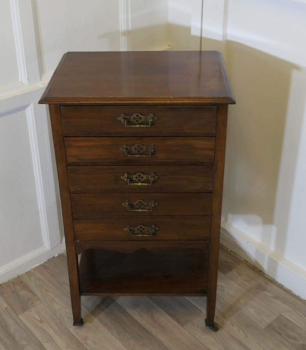 An Arts and Crafts warm brown wood music cabinet with five drawers featuring ornamental brass drop handles, raised on tapered legs with castors and a lower shelf. View III