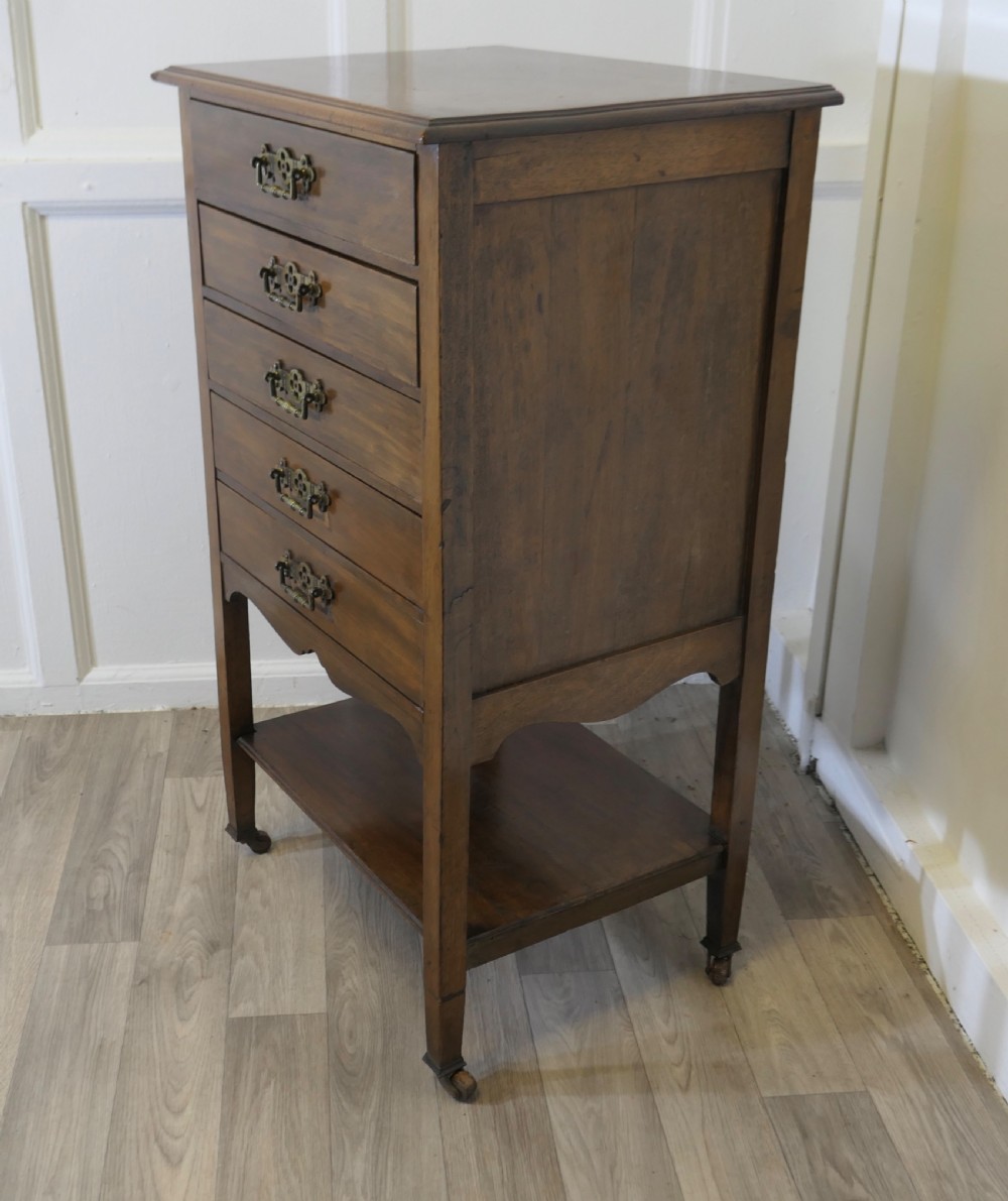 An Arts and Crafts warm brown wood music cabinet with five drawers featuring ornamental brass drop handles, raised on tapered legs with castors and a lower shelf. View V