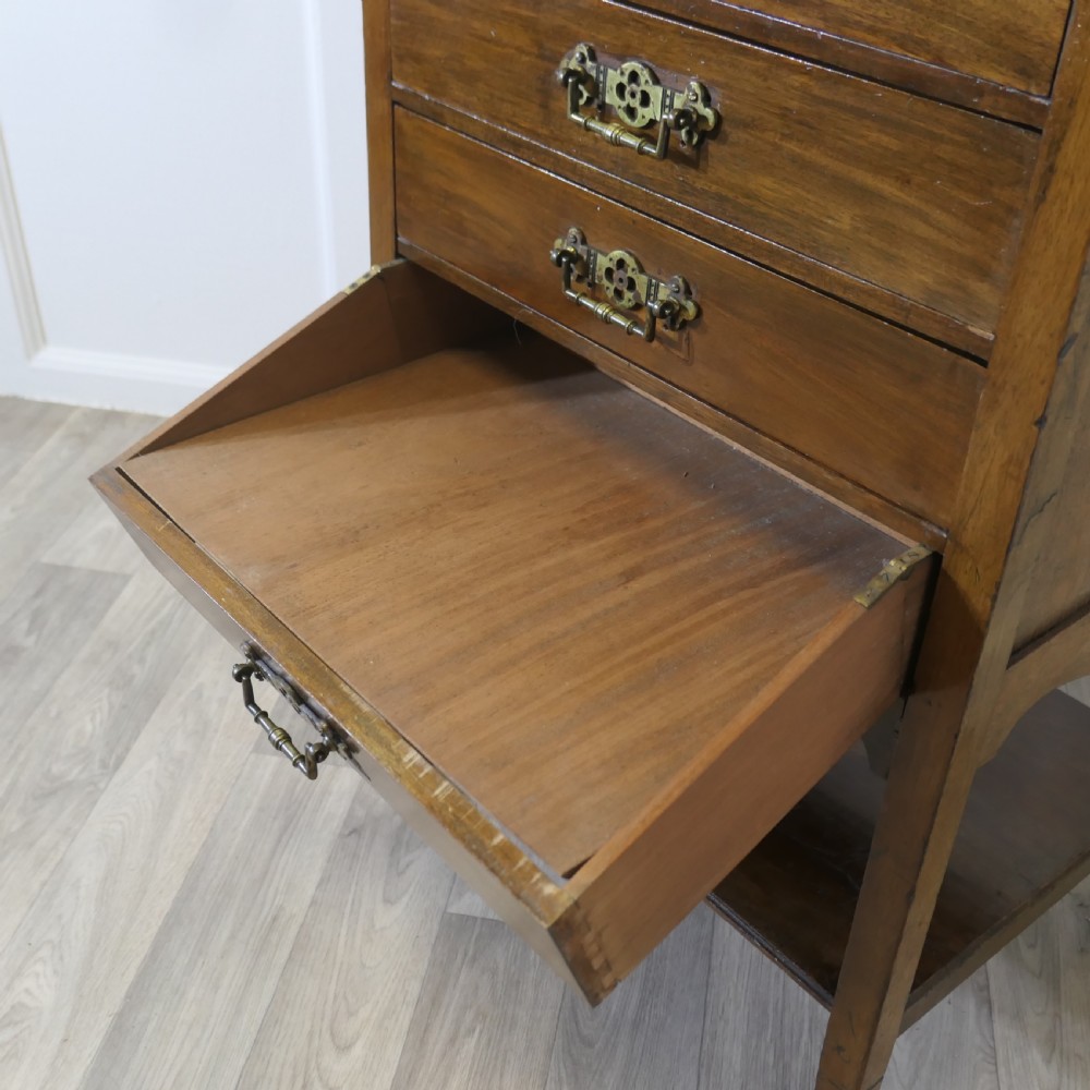 An Arts and Crafts warm brown wood music cabinet with five drawers featuring ornamental brass drop handles, raised on tapered legs with castors and a lower shelf. View VII