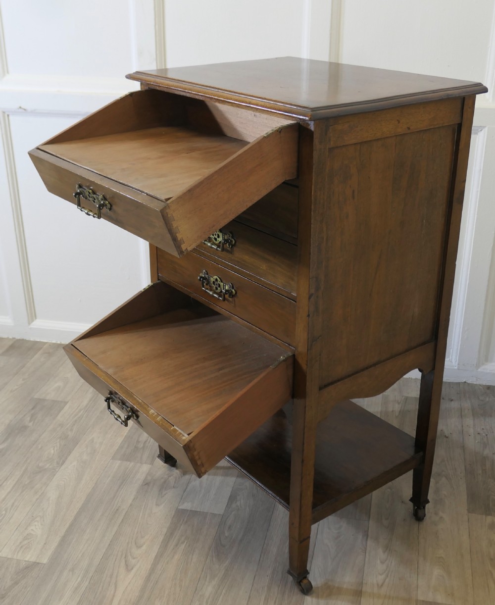 An Arts and Crafts warm brown wood music cabinet with five drawers featuring ornamental brass drop handles, raised on tapered legs with castors and a lower shelf. View VIII