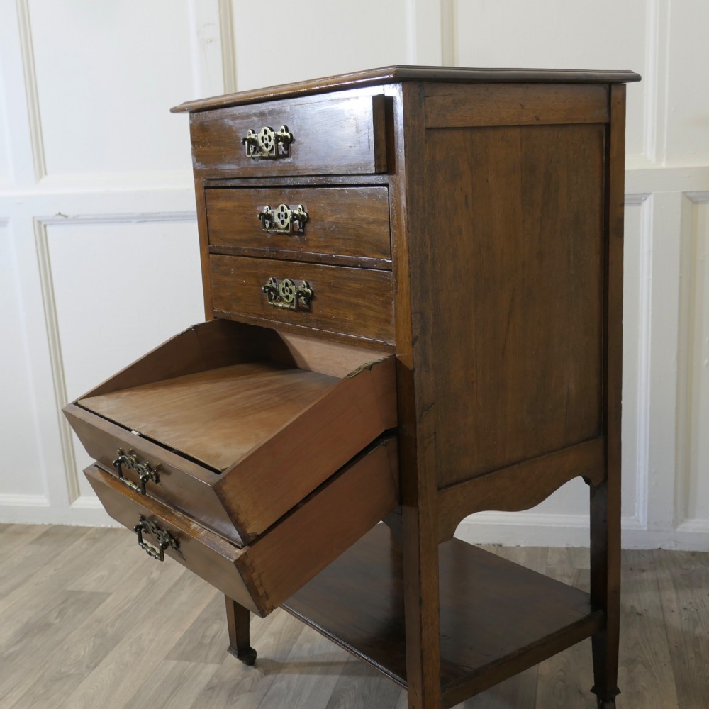 An Arts and Crafts warm brown wood music cabinet with five drawers featuring ornamental brass drop handles, raised on tapered legs with castors and a lower shelf. View X