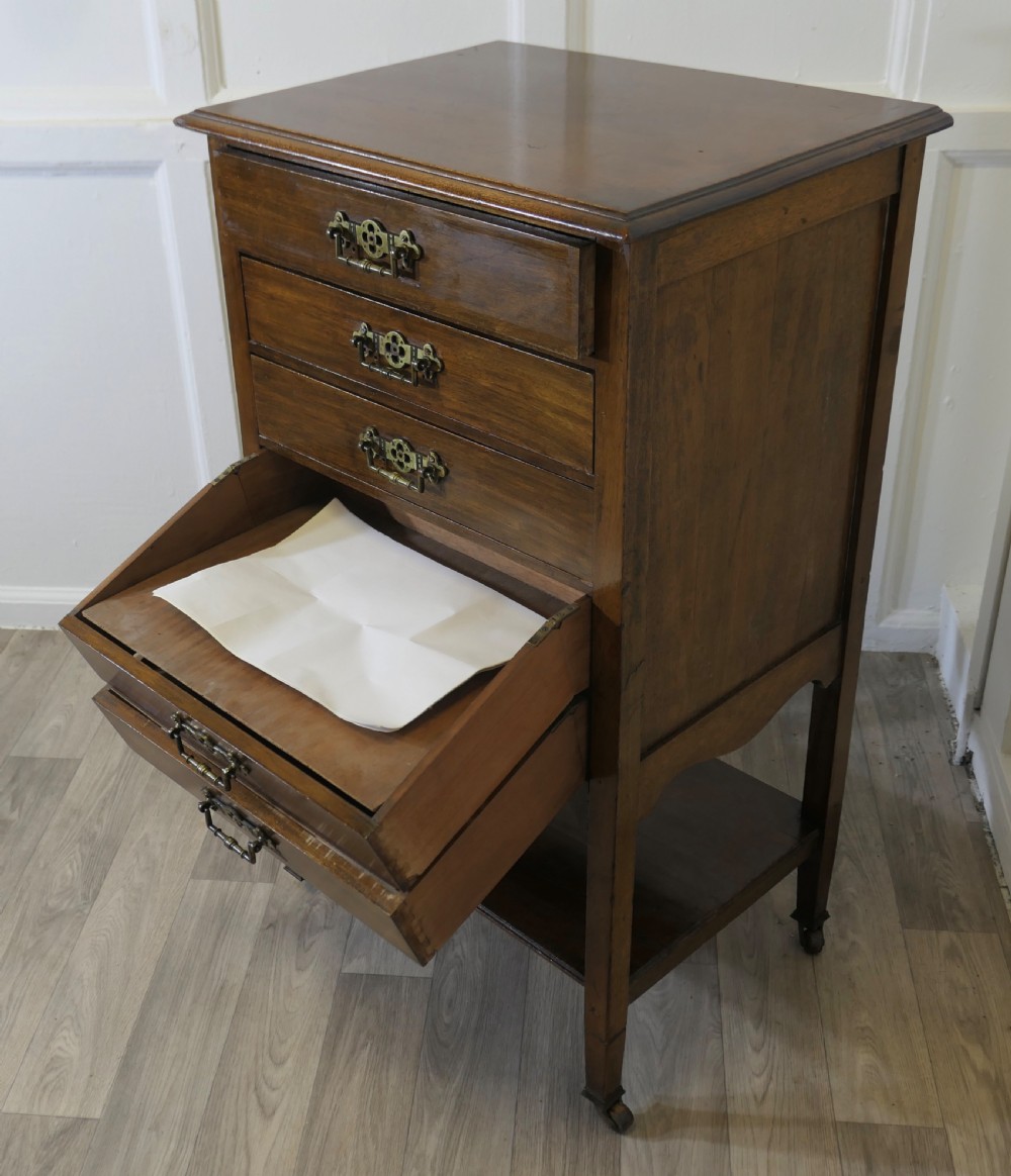 An Arts and Crafts warm brown wood music cabinet with five drawers featuring ornamental brass drop handles, raised on tapered legs with castors and a lower shelf. View XI