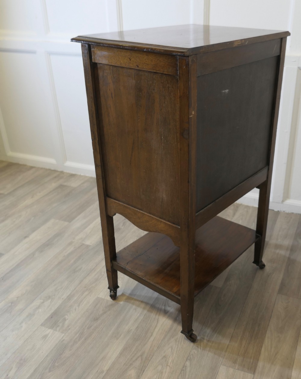 An Arts and Crafts warm brown wood music cabinet with five drawers featuring ornamental brass drop handles, raised on tapered legs with castors and a lower shelf. View XII