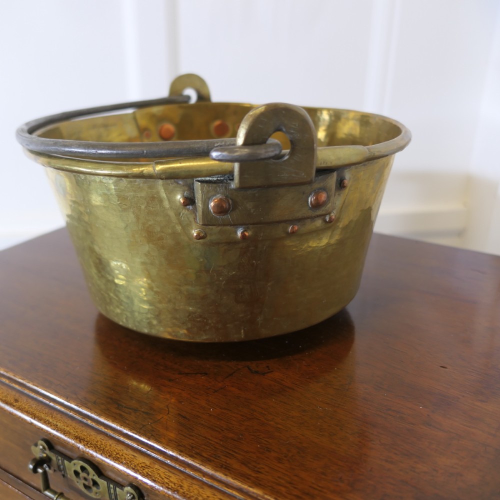 A small 19th-century hand-hammered brass pan with copper rivets and an iron swing handle resting on a polished wooden surface.