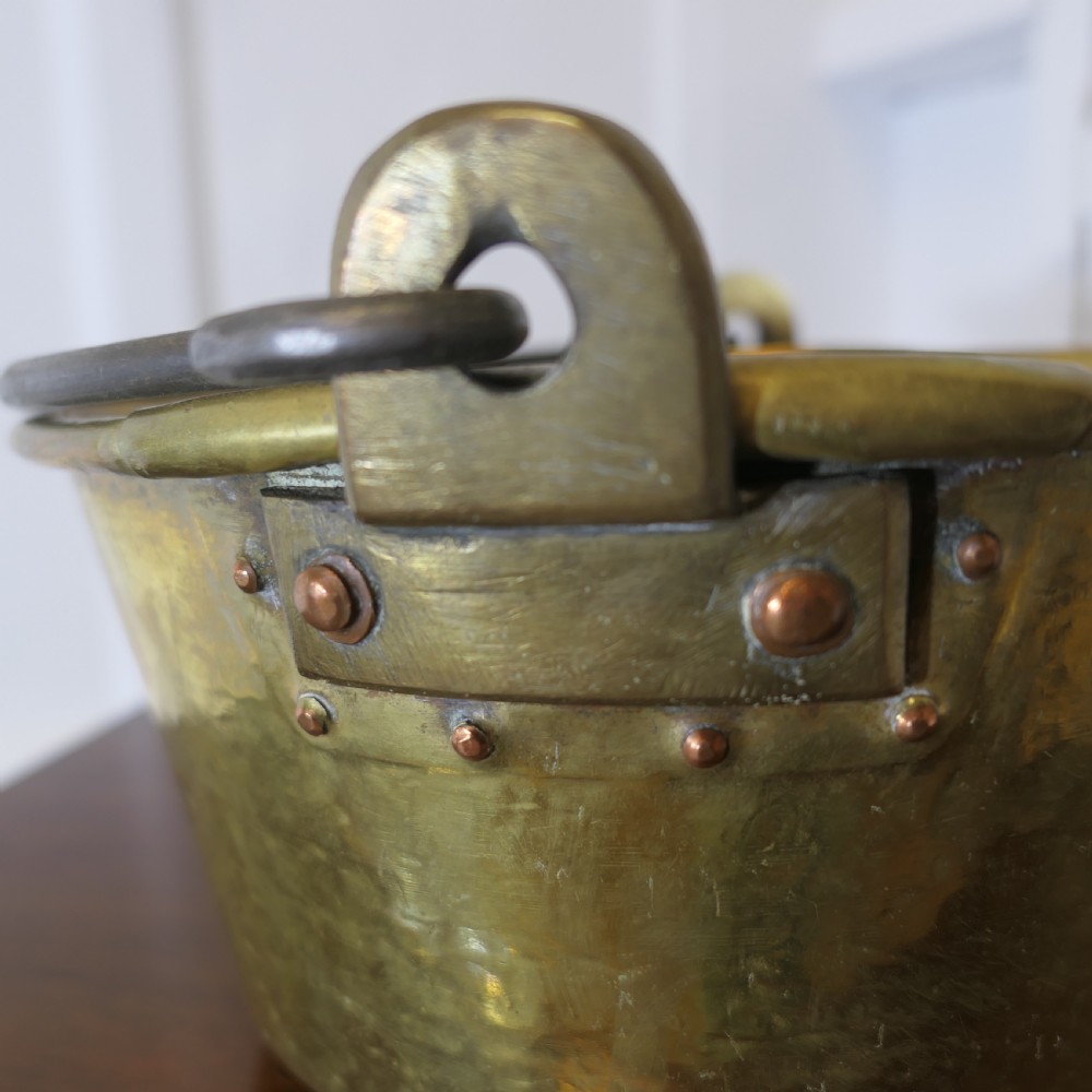 A small 19th-century hand-hammered brass pan with copper rivets and an iron swing handle resting on a polished wooden surface. View II