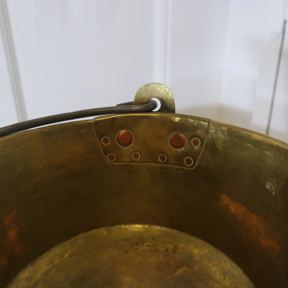 A small 19th-century hand-hammered brass pan with copper rivets and an iron swing handle resting on a polished wooden surface. View III