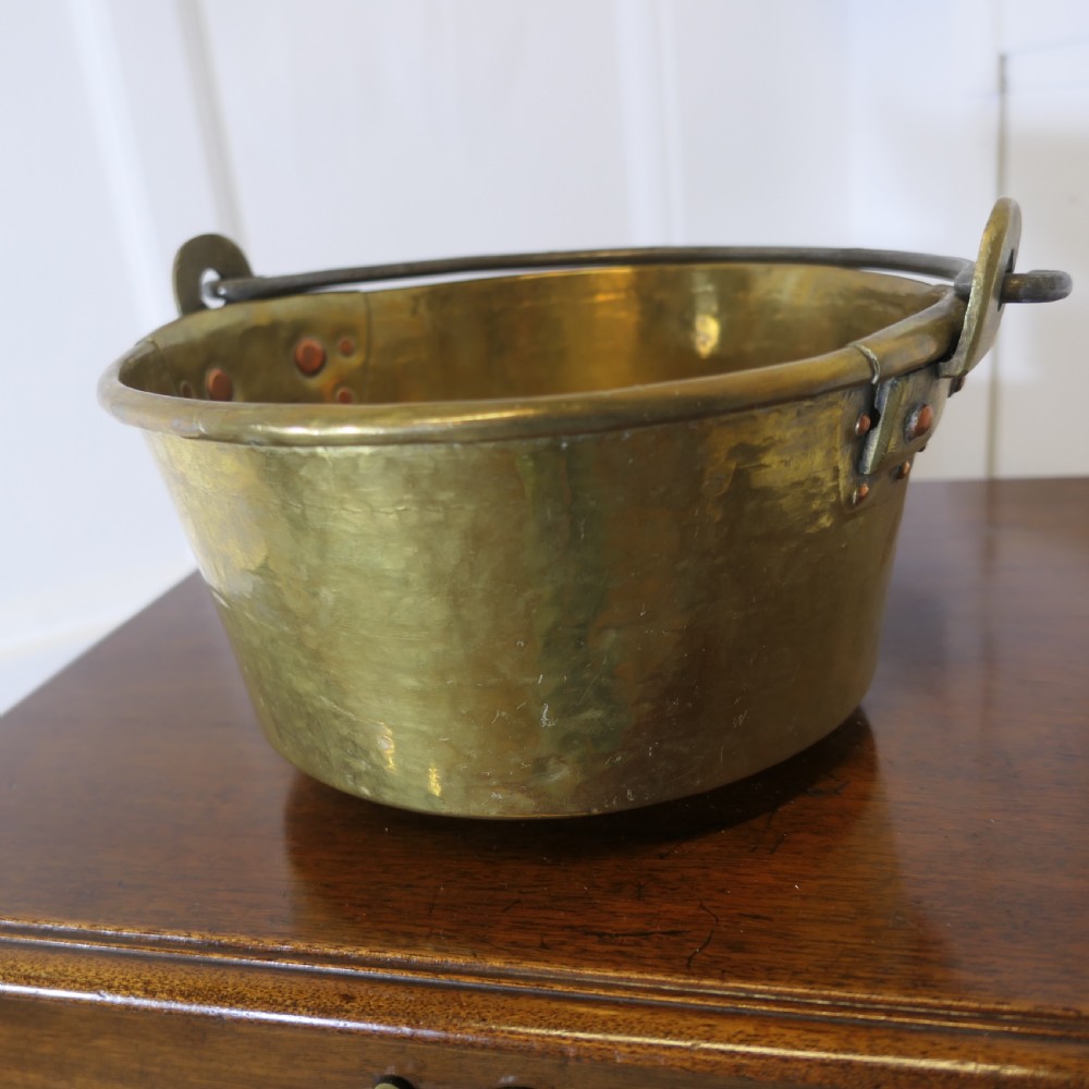 A small 19th-century hand-hammered brass pan with copper rivets and an iron swing handle resting on a polished wooden surface. View IV