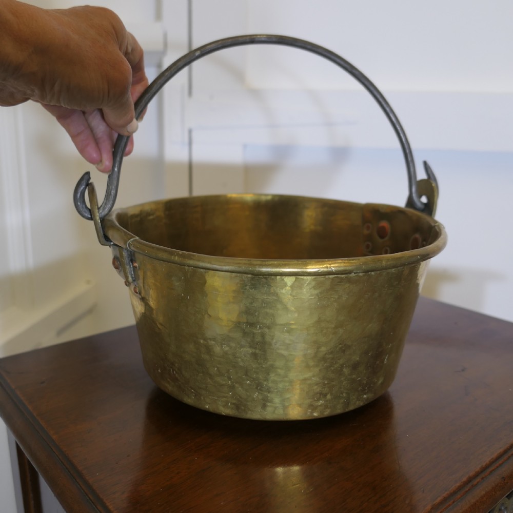 A small 19th-century hand-hammered brass pan with copper rivets and an iron swing handle resting on a polished wooden surface. View V
