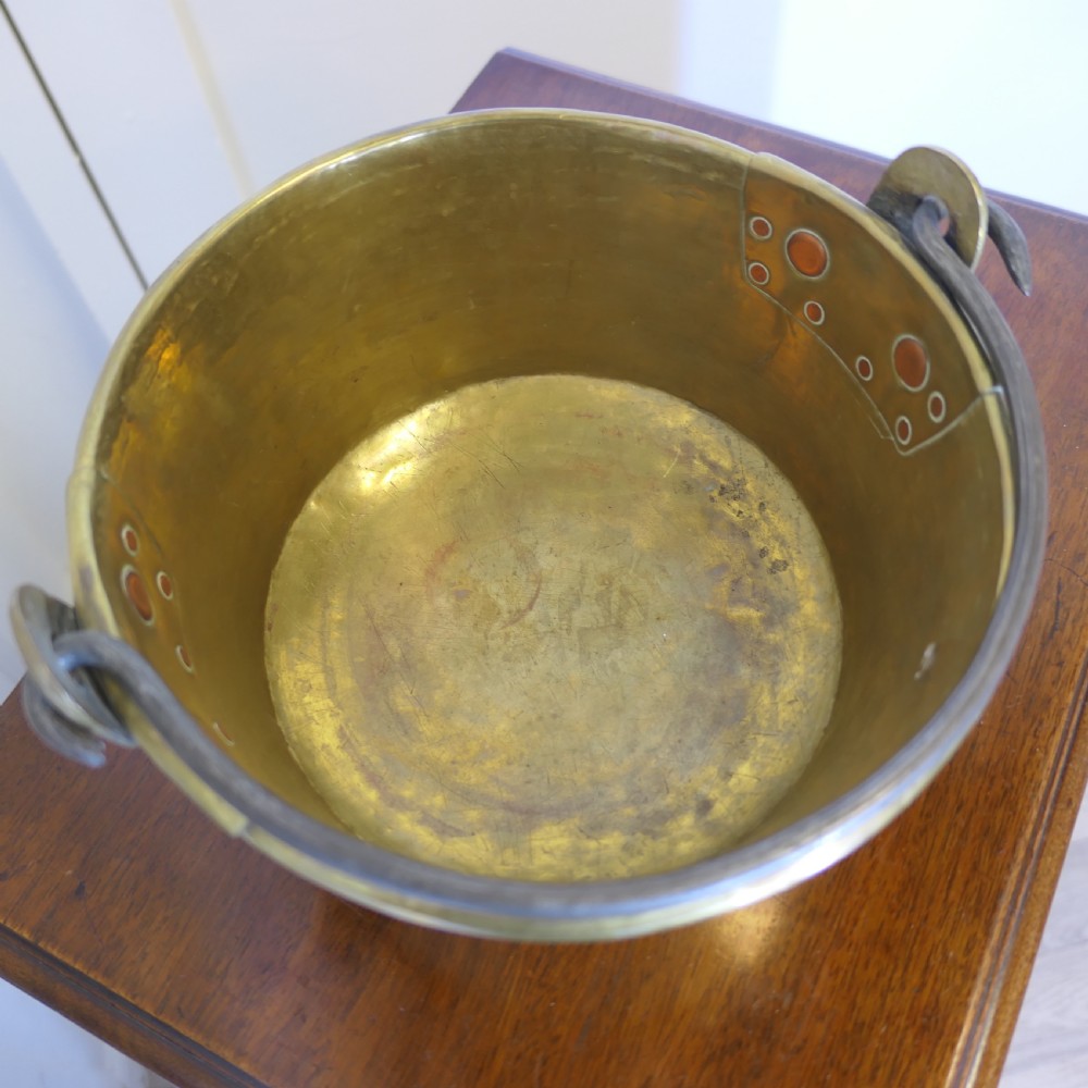 A small 19th-century hand-hammered brass pan with copper rivets and an iron swing handle resting on a polished wooden surface. View VI