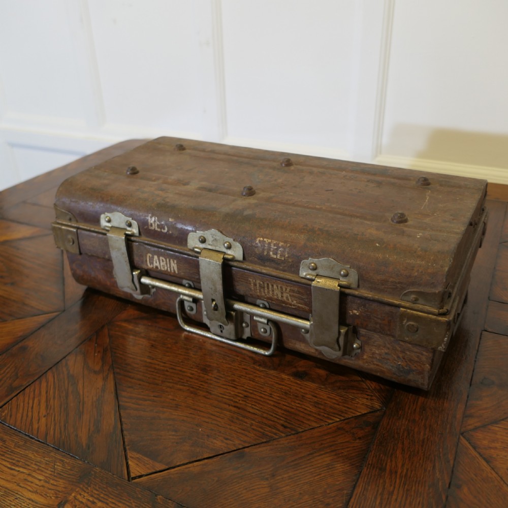 A small dark brown steel cabin trunk featuring three front metal latches, a central folding handle, and painted lettering reading Best Steel Cabin Trunk.