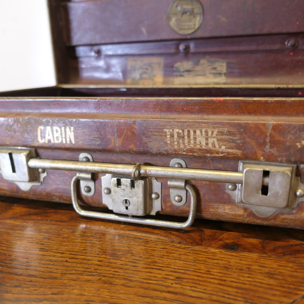 A small dark brown steel cabin trunk featuring three front metal latches, a central folding handle, and painted lettering reading Best Steel Cabin Trunk. View VII