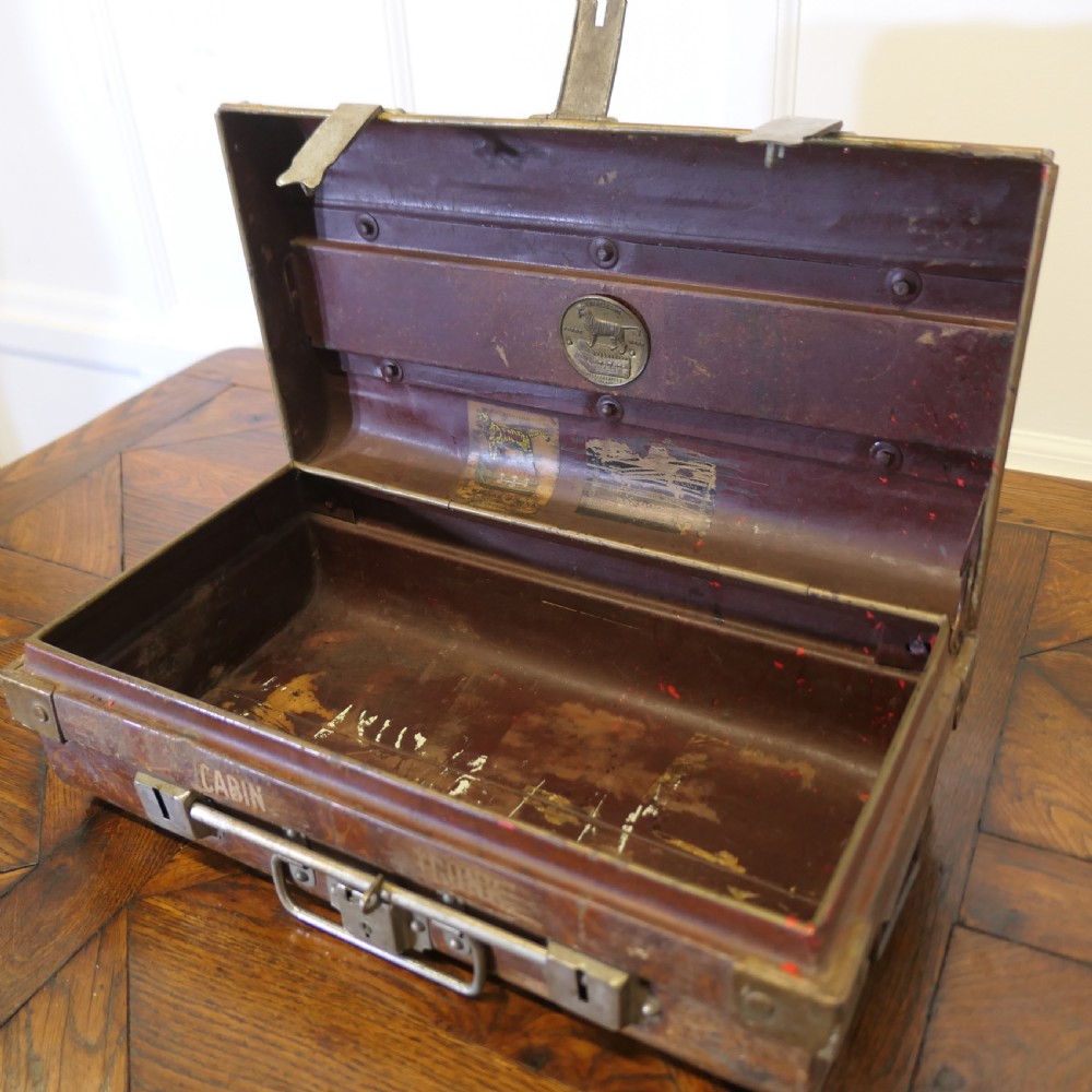 A small dark brown steel cabin trunk featuring three front metal latches, a central folding handle, and painted lettering reading Best Steel Cabin Trunk. View VIII