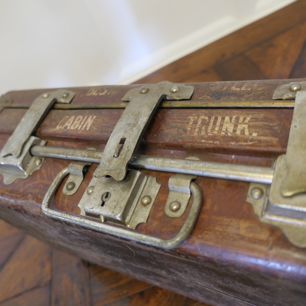 A small dark brown steel cabin trunk featuring three front metal latches, a central folding handle, and painted lettering reading Best Steel Cabin Trunk. View X