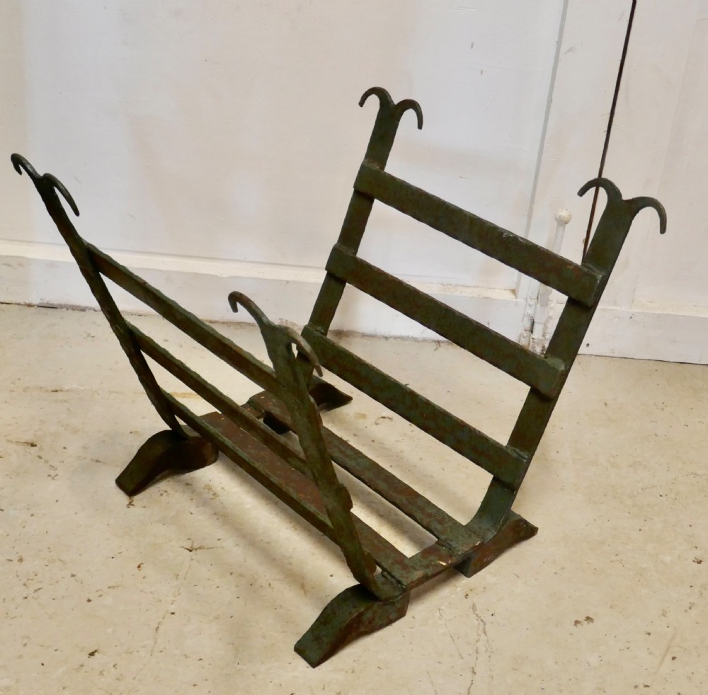 A heavy hand-forged wrought iron log carrier with angled side slats, scrolled finials, and a patinated green and rust finish.