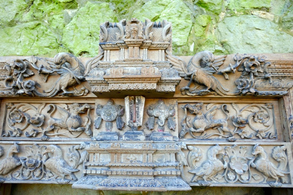 An intricately carved natural teak wooden archway featuring relief carvings of elephants and architectural motifs. View V