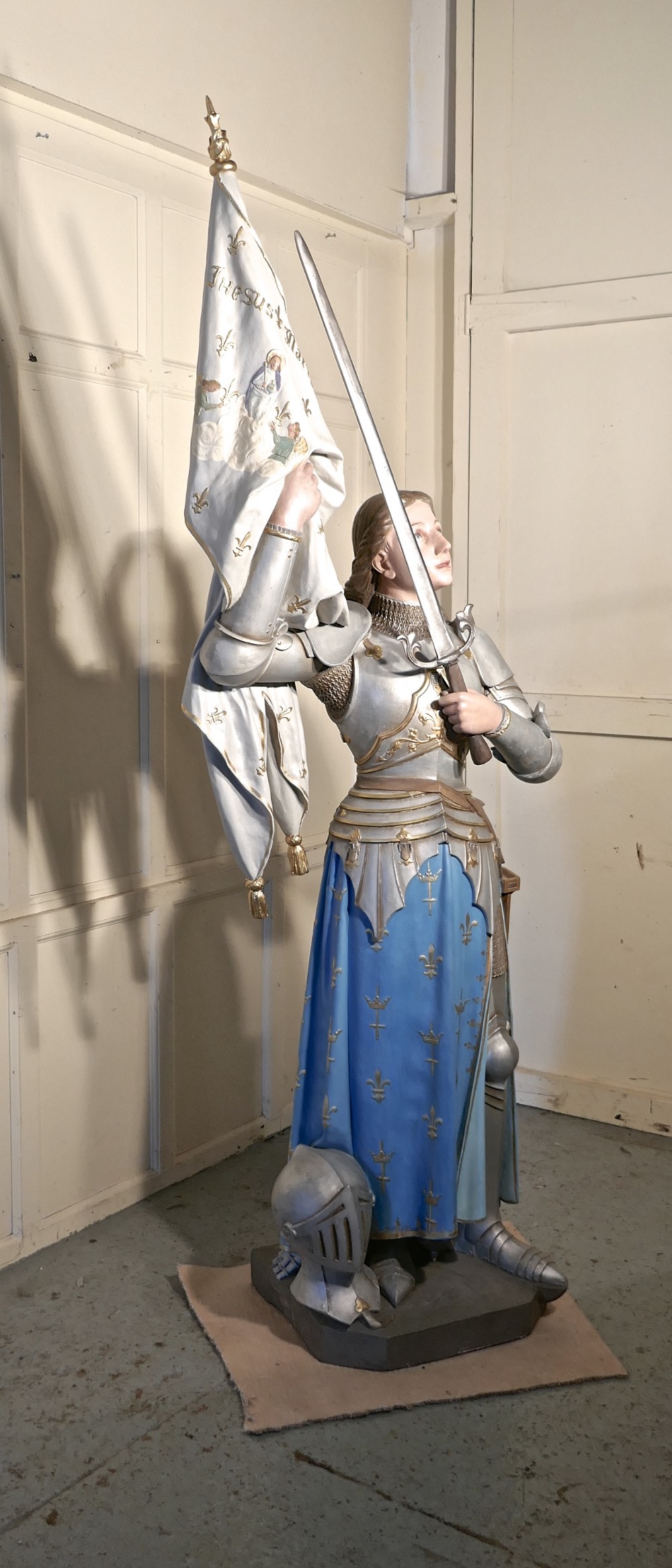 A French polychrome plaster statue depicting Saint Joan of Arc in silver armour and a blue tunic decorated with fleur-de-lis, holding a sword and flag.